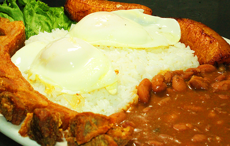 Plate of fried pork, white rice topped with fried eggs, stewed beans, fried plantains, and lettuce garnish; warm tones on a white plate