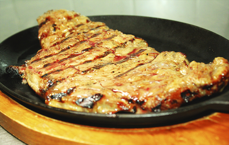 Grilled steak with char marks served on a black sizzling platter atop a wooden board; dark brown and charred tones