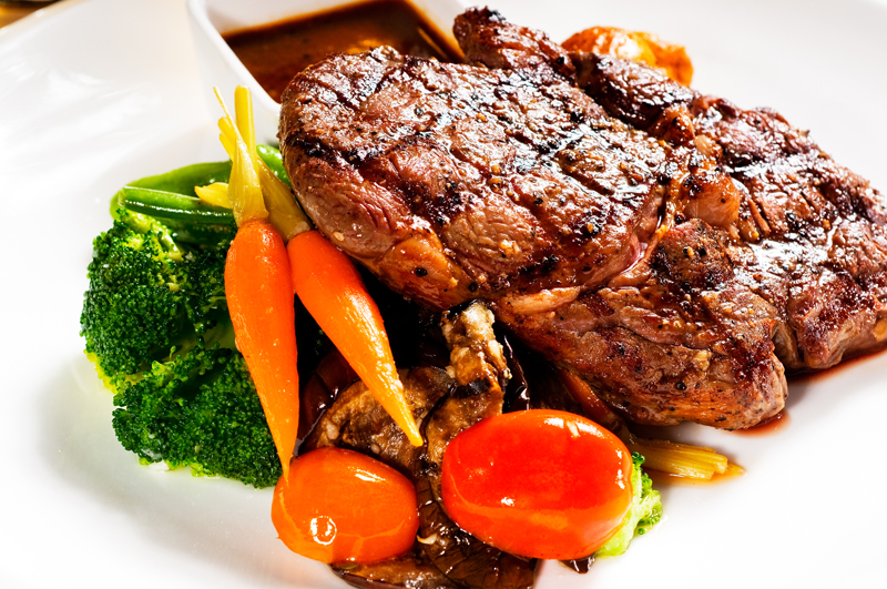 Grilled steak plated with broccoli, baby carrots, mushrooms, cherry tomatoes and a side of sauce